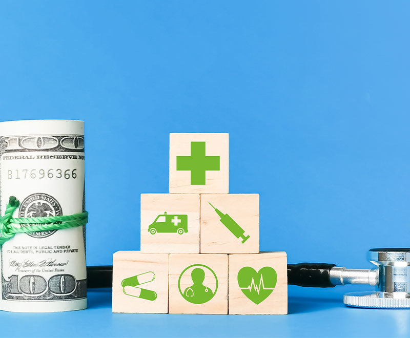 Photo of rolled up dollar bills, stethoscope and building blocks with healthcare icons on them.