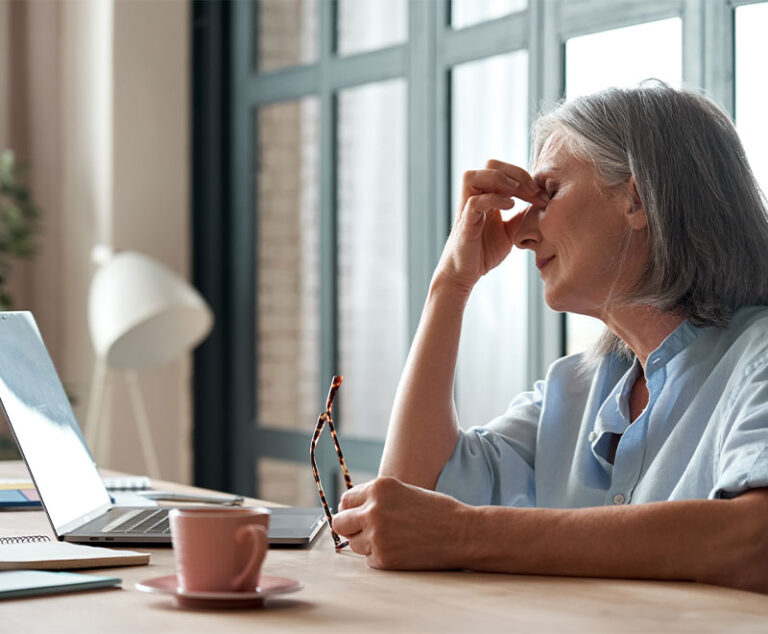 Photo of a senior woman pinching the bridge of her nose while sitting at her computer