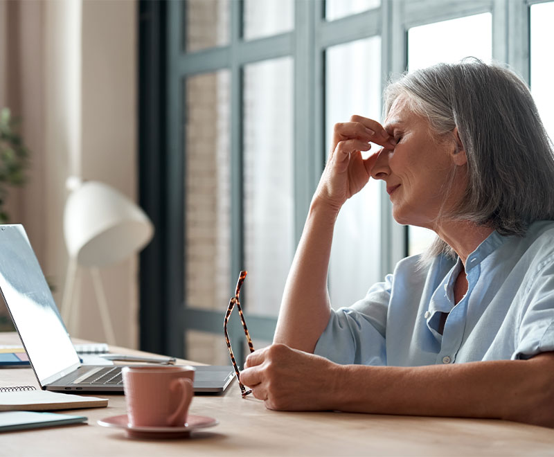 Photo of a senior woman pinching the bridge of her nose while sitting at her computer