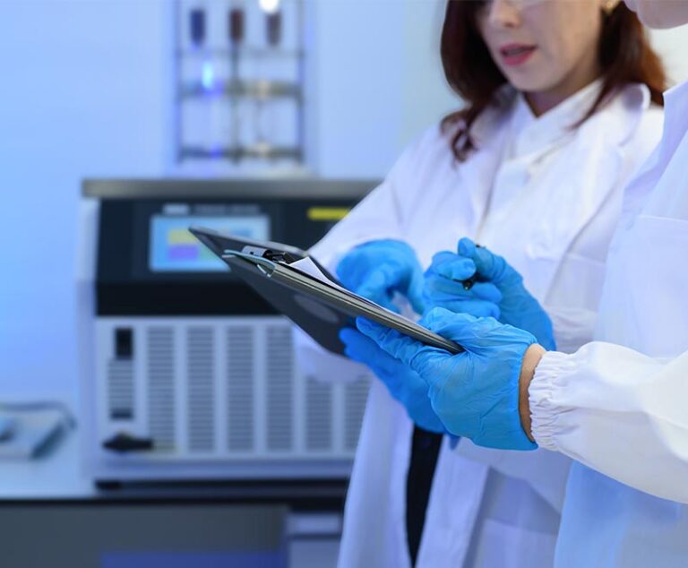 Two scientists in a lab wearing blue gloves and holding clipboards.