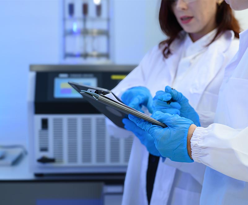 Two scientists in a lab wearing blue gloves and holding clipboards.